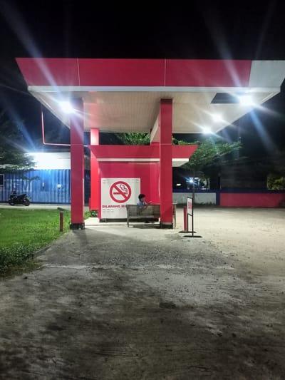 An outdoor scene at night depicts a covered area, likely a bus stop or rest area, with bright overhead lighting illuminating the space. The structure is characterized by a red roof with white undersides, supported by red pillars. In the center, a "No Smoking" sign is prominently displayed on a red wall, with text below it reading "DILARANG MEROKOK" which translates to "NO SMOKING". To the right of the sign, a young person is seated on a wooden bench, looking down at a mobile phone. Adjacent to the covered area, a blue shipping container is visible, and a black motorcycle is parked near it. The ground is a mixture of concrete and gravel. A sign with the word "BUKA" meaning "OPEN" is also present. Trees are visible in the background, and the overall environment suggests a location within the city of Tanjongkeumala, Indonesia.
