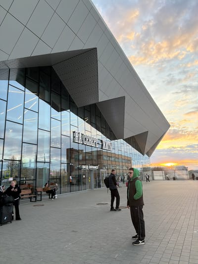 The image shows a modern building with glass windows and gray cladding in Murmashi, Russia. The building's exterior features sharp geometric angles. Two people are walking on the sidewalk in front of the building. Other people are sitting on benches. The sky displays colors of a sunset or sunrise. The word "ENTRANCE" is visible on the building's facade.