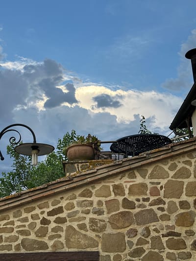 The image shows a stone wall with a tiled roof, a chimney, and a balcony with a table and chairs. There is also a potted plant and a lamp post. The sky is blue with some clouds. The scene appears to be in Cannara, Italy, a town known for its historic buildings and natural beauty. The stone wall is made of irregular shaped stones, some of which are covered in moss. The tiled roof is made of terracotta tiles. The chimney is black and made of brick. The balcony is furnished with a black metal table and chairs, and a potted plant containing succulents. The lamp post is black with a curly arm and a round lampshade. The sky is a bright blue with scattered white and grey clouds.