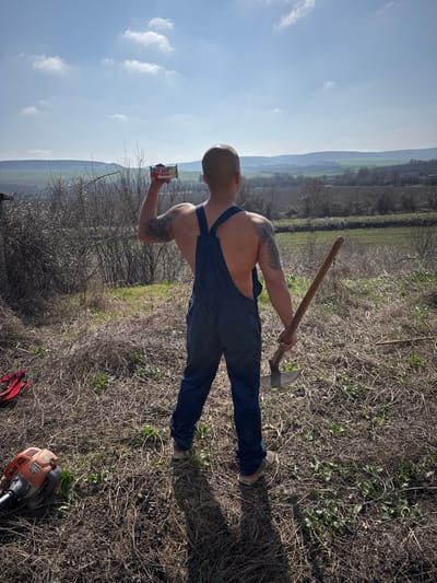 An outdoor scene in Katselovo, Bulgaria, depicts a male individual viewed from behind, standing on dry, matted ground under a bright blue sky with scattered clouds. The man is bare-chested, wearing dark blue denim overalls, and exhibits tattoos on both upper arms. He holds a red, white, and blue packaged snack bar, labeled "BATE CHOCO," aloft in his raised right hand. His left hand grips a long-handled tool, likely an axe or mattock, with the implement's head resting on the ground. To the lower left, an orange-and-white gas-powered string trimmer lies on the ground, adjacent to a crumpled red item. The surrounding environment consists of sparse vegetation, bare trees and shrubs in the mid-ground, and cultivated fields leading to distant rolling hills under the clear sky. Shadows indicate a mid-morning or late afternoon sun position.
