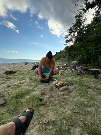 A person with dark hair pulled into a bun, wearing a yellow t-shirt and blue shorts, squats on a sandy beach in Mgudzyrkhwa, Abkhazia, near the water. The person is preparing food on a grill, which is placed on the sand next to some rocks. The beach is covered in sand with patches of green grass. In the background, there is a body of water, a line of trees, and a cloudy sky. In the foreground, a bare foot wearing a black sandal is visible.