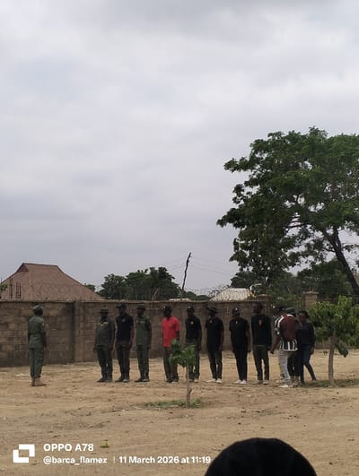 The scene is set outdoors on an unpaved, dry, reddish-brown dirt ground in Gidan Kado, Nigeria, under an overcast sky. In the mid-ground, one individual in olive-green military-style fatigues, including a cap and boots, stands facing right, addressing a line of eleven other individuals. Their back is partially visible to the viewer.
The line of eleven individuals stands across the mid-ground, facing the single individual. From left to right:
* The first two individuals in the line wear olive-green uniforms with black caps.
* The next five individuals wear black shirts, dark trousers, and black caps; one of these (the third from the left in this black-clad group) has a red long-sleeved shirt visible beneath his black shirt.
* The eighth individual in the line wears a red short-sleeved shirt, dark trousers, and a black cap.
* The final three individuals on the right of the line include two men and two women. One man wears a light-colored striped shirt under an open dark jacket, and another wears a dark top and trousers. One woman wears a black long-sleeved shirt and blue jeans, and the other wears a black top and dark trousers. All individuals in the line are standing upright in a formation.
In the background, a low, light-brown brick wall topped with barbed wire runs horizontally. Behind the wall, various residential structures with light-brown corrugated metal roofs are visible, interspersed with green trees and bushes. A large tree with dense green foliage occupies the right side of the background. Two small saplings are planted in the dirt ground in the mid-foreground. A dark, out-of-focus object, possibly a person's head, is visible in the bottom right corner of the frame.