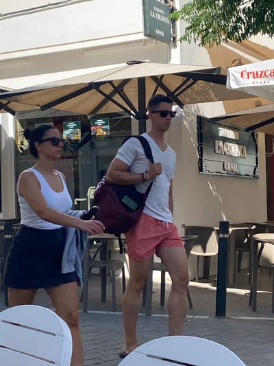Two people, a woman and a man, are walking on a sidewalk in front of a building with an awning and umbrellas. The woman, on the left, is wearing sunglasses, a white tank top, and a dark skirt. She has her dark hair pulled back in a bun and is carrying a light-colored garment over her left arm. The man, on the right, is wearing sunglasses, a white t-shirt, and red patterned shorts. He has a dark bag slung over his right shoulder. Behind them, a sign for "TABERNA LA CHANCICA CONIL" is visible on the building. Tables and chairs for an outdoor seating area are set up in front of the establishment. The street is paved with cobblestones. The lighting suggests it is daytime, likely a sunny day given the shadows.