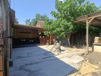 This image depicts an outdoor, rural scene in Peremozhne, Ukraine, likely during the daytime given the bright sunlight and clear blue sky. The setting appears to be a yard or courtyard with elements of construction or renovation in progress.

The main subjects include a concrete mixer, which is centrally placed on the paved ground. A long, narrow wooden plank lies horizontally in the foreground, possibly for mixing or leveling concrete. The ground itself is a mix of concrete and dirt, with some areas appearing freshly poured and others more weathered.

In the background, there is a wooden structure with a sloped, corrugated roof, possibly a shed or a covered patio. This structure has vertical wooden planks for its walls and a wooden deck area with benches. Several white potted plants hang from the structure, adding a touch of greenery. To the right, a large tree with broad green leaves is prominent, casting shadows on the scene. Further to the right, a covered area with a wooden frame and a corrugated roof is visible, with stacks of bagged material (likely cement or similar construction supplies) piled next to it. A wire mesh fence is also visible in the background, suggesting boundaries or enclosures.

The overall impression is one of outdoor work or DIY projects. There are no people visible in the image. The weather appears to be sunny and warm. The presence of the concrete mixer, unfinished concrete areas, and building materials strongly indicates an ongoing construction or repair activity. The scene is utilitarian and functional, with the natural elements of the tree and sky contrasting with the man-made structures and materials. No visible text is present in the image.