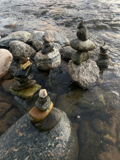 A circle of balanced rocks sits in a shallow stream. Water flows around and over the rocks, creating ripples and reflections. The stones vary in size, shape, and color, from smooth gray pebbles to larger, rougher rocks with green algae. The composition is natural and peaceful, evoking a sense of serenity and balance. The focus is on the harmony between the rocks and the water.