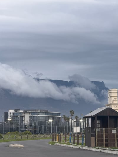 The image depicts Table Mountain partially obscured by clouds, viewed from Milnerton, South Africa. In the foreground, a road with a concrete barrier runs along the right side. Adjacent to the road is a grassy area with sparse vegetation. Buildings, including a tall, modern structure with multiple windows, are visible behind the grass. There is a wooden structure and a white cylindrical building in the middle ground. Palm trees line part of the street. The sky is overcast with varying shades of gray.