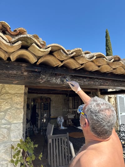 An outdoor scene depicts a man wearing safety glasses, a face mask, and a glove, working on a wooden post or beam next to a roof made of curved tiles. The man appears to be cleaning or examining the post with a small tool in his gloved hand. The post is dark in color and shows signs of aging or wear. The setting seems to be in Tourrettes-sur-Loup, France, indicated by the architectural style with stone buildings and the presence of a cypress tree in the background under a clear blue sky. The scene may be taking place at a restaurant.