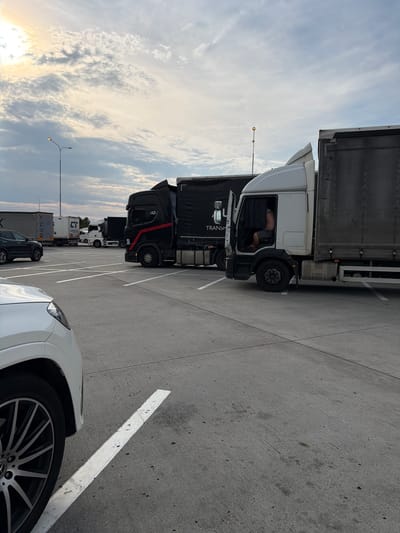 The image depicts a parking lot filled with various trucks and a white car. In the foreground, the front passenger side of a white SUV is visible, showcasing its alloy wheel. Behind it, a line of parked trucks stretches into the distance. The trucks are of different colors, including black and white, and appear to be commercial vehicles. Some have trailers attached, with visible branding.
The scene is set on a paved surface, likely a parking lot, with distinct white lines marking parking spaces. The sky above is partly cloudy with streaks of sunlight suggesting either sunrise or sunset. A streetlamp is visible in the distance. A person can be seen in the driver's cabin of a white truck, appearing to be resting or taking a break.
The image is oriented vertically, with the sky at the top and the ground at the bottom, but the content is rotated 90 degrees counter-clockwise. This suggests the photograph was taken in landscape orientation and then rotated.
There is some visible text on the trucks, including "TRANS", "MAN", and "BENIX". The overall impression is a quiet moment in a commercial transport hub, possibly during a rest stop, with the natural light indicating a transitionary time of day. The clouds and sunlight create a soft, diffused lighting. The location context of Křenovice, Czechia, places this scene within a specific geographical area, though no explicit landmarks are visible.