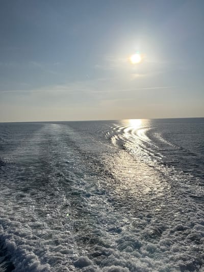 The image depicts a vast expanse of open sea under a bright, clear sky, viewed from the stern of a moving vessel in the waters near Kamariotissa, Greece. A prominent, frothy white wake originates from the immediate foreground, extending in a relatively straight path towards the distant horizon, indicating the vessel's propulsion. The sky is light blue with sparse, faint cirrus clouds and two visible linear contrails from aircraft. The sun, appearing as a brilliant white orb with a surrounding halo, is positioned in the upper right quadrant of the sky. Its intense light creates a shimmering, reflective path across the water's surface, extending from the horizon towards the viewer, intersecting with the vessel's wake. The sea itself is a deep blue-grey, appearing darker in areas not directly illuminated by the sun's reflection. No individuals or specific man-made structures are discernible within the frame, aside from the implied vessel and atmospheric contrails.