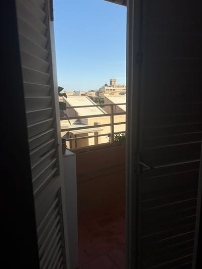The image captures a view through an open doorway and window with louvred shutters, revealing a cityscape under a clear blue sky. The interior foreground is dominated by the dark, shadowed interior of the room and the texture of the louvred shutters on both the left and right sides of the frame. To the left, a white, vertical structure with a textured surface is visible.

The exterior view shows a tiled balcony or terrace with a reddish-brown floor. Beyond this, there is a tan railing and a building with a flat, light-colored roof. Further in the distance, a cluster of buildings with terracotta roofs and light-colored facades are visible, with a prominent tower structure featuring a clock face and a spire. The architecture suggests a European setting, possibly in a Mediterranean climate. The scene is devoid of any people. The image appears to be taken during daylight hours, as indicated by the bright sky and the clear visibility of the external environment. The perspective suggests the viewer is looking out from a building.
