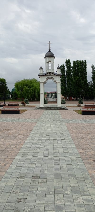 The image depicts a paved public square in Kirsanov, Russia. In the center stands a white ornate structure resembling an archway topped with a dome and a cross, suggesting a religious or memorial significance. Through the archway, a fountain with multiple jets of water is visible. The square is paved with two-toned bricks, arranged in a patterned walkway leading towards the central structure. To the sides of the walkway, there are benches and some greenery, including trees and bushes. The sky is overcast, casting a soft, diffused light over the scene. The overall impression is that of a serene and well-maintained public space.