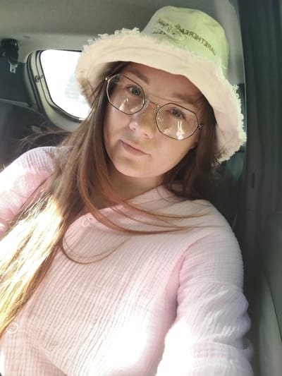 A young woman with long, light brown hair and freckles is taking a selfie in a car. She is wearing a pale pink, crinkled shirt with buttons and a light-colored bucket hat. She also has on round, wire-rimmed glasses. The setting appears to be the interior of a vehicle, with a window visible behind her and the dark interior of the car in the foreground and right side. Sunlight streams in from the left, illuminating her hair and the left side of her face and shirt. The mood seems relaxed and casual, possibly indicating a daytime outing. There is some text visible on the bucket hat, but it is not legible. The location context is Grabovo, Russia.