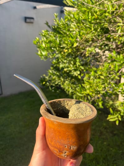 A hand holds a gourd filled with yerba mate and a metal straw, likely outdoors. The location is speculated to be Gerli, Argentina. A blurred background shows foliage and a building, suggesting a residential or garden setting. The gourd's contents appear dry and crumbly. The scene is brightly lit, indicating daytime.