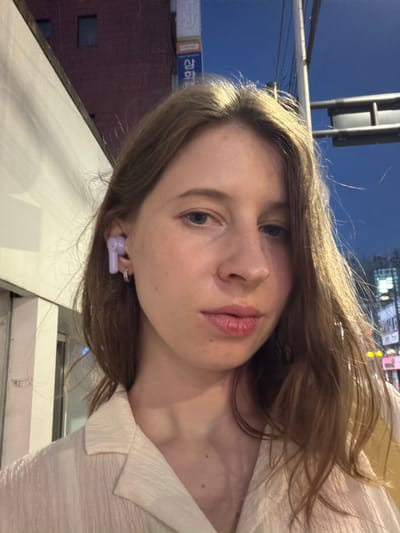 A young Caucasian woman is taking a selfie outdoors at dusk. She has long, light brown hair that is slightly windswept and is wearing a light-colored, textured collared shirt. She has a purple earbud in her right ear and a small hoop earring in her earlobe. Her lips are slightly parted. In the background, there is a building with a dark red facade and two dark rectangular windows. Above the building, there is a blue sky with a street light fixture visible. Signs with Korean text are also visible in the background, suggesting the location is Uijeongbu-si, South Korea. The lighting suggests it is twilight.