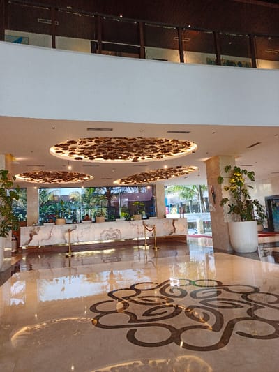 The image showcases a hotel lobby with a high ceiling featuring three large, circular light fixtures with brown patterns. Below, a reception desk with staff overlooks a bright outdoor view through large windows. Two large potted plants flank the lobby entrance. The polished marble floor displays a decorative swirling pattern. The upper level of the lobby with a dark-toned railing is visible in the background.