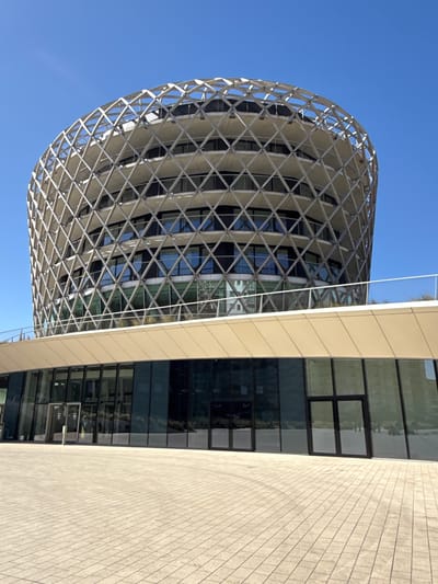 A modern architectural scene in Middelkerke, Belgium, is captured on a clear, sunny day. The image features two distinct but connected building structures under a bright blue sky.
On the left, a prominent spherical or dome-like building dominates the view. Its exterior is encased in an intricate, light-colored lattice or diagrid structure, forming a network of diamond and square shapes. This lattice appears to be made of metal, possibly white or silver, and covers a facade of dark, reflective glass that mirrors the sky and surrounding elements.
To the right of the spherical structure, a more rectilinear building stands, characterized by a smooth, light beige or cream-colored facade. This section features multiple large, dark-tinted rectangular windows, which also reflect the clear blue sky and the complex geometry of the adjacent spherical building.
The foreground on the bottom right reveals a paved surface, consisting of light-colored, rectangular paving stones or tiles, suggesting a plaza or walkway area. There are no people or discernible activities taking place, and no visible text is present. The overall impression is one of modern design and architectural innovation, highlighted by the strong natural light of a cloudless day.