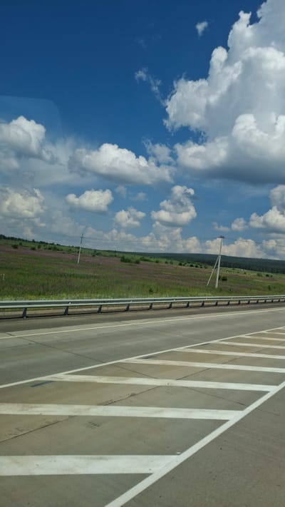 The image shows a highway in Kansk, Russia on a sunny day with a blue sky and white clouds. The highway has multiple lanes and white markings. On the side of the highway, there is a grassy field with purple flowers and trees. There are also utility poles visible in the distance. The image appears to be taken from a moving vehicle, as indicated by the slight blur and the perspective. There are no people visible in the image.