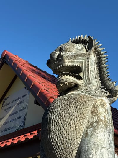 The image shows a stone statue of a mythical creature, possibly a lion or a dragon, positioned in front of a building with a red tiled roof. The statue is detailed, with a fierce expression, sharp teeth, and a mane of spikes. The building in the background has a sign with Thai writing. The sky is clear and blue. This scene is located in Baan Tha Po, Thailand.