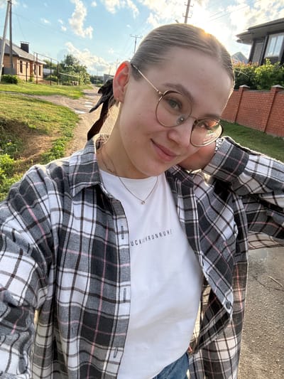 This is a vertical, close-up selfie of a young woman taken outdoors. The woman has blonde hair pulled back into two braids and is wearing glasses with thin, gold-colored frames. She is dressed in a white t-shirt with black text on the chest and a gray, black, and pink plaid open-front shirt over it. The text on her t-shirt appears to be in German, reading "KEHRUNRUHE". She is looking at the camera with a slight smile.
The background suggests a rural or suburban setting. There are houses with brick and dark siding visible, along with green grass and trees. A dirt or gravel path runs through the scene. The sky is blue with scattered white clouds, and the sun is low in the sky, casting a warm glow and creating a lens flare on the left side of the image. This indicates it is either early morning or late afternoon. The lighting suggests the weather is clear and pleasant. The woman's relaxed pose and smile convey a casual and happy mood.