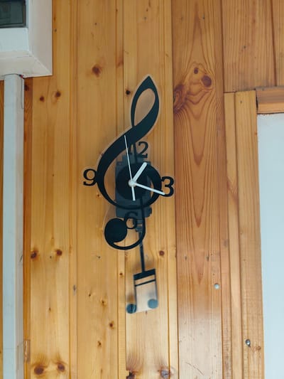A musical note-shaped clock is mounted on a light-colored wooden paneled wall. The clock has black hands and numerals. The hour hand points to the 9, and the minute hand points to the 12. A small musical note pendulum hangs below the clock. The wall appears to be an interior wall, likely in a home or a business. There is a white frame on the left side of the image, and a white wall on the right side. The image is a close-up shot, focusing on the clock and the wall. No people are visible in the image. The scene depicts a decorative clock on a wooden wall, located in Edineț, Moldova.