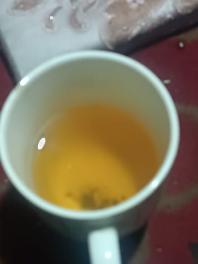 A top-down shot shows a white mug filled with a clear, amber-colored liquid, possibly tea, with a dark clump of leaves or tea bag visible at the bottom. The mug sits on a surface with hints of red and possibly black. A blurry, out-of-focus object rests behind the mug and contains light and dark elements, potentially a book or decorative tray. A subtle highlight is visible on the liquid's surface.