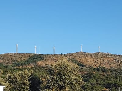 The image depicts a view of a hilly landscape with wind turbines on the horizon. The sky is clear and blue. In the foreground, there are trees and foliage. The middle ground shows a rocky, arid hillside dotted with vegetation. The background features a line of wind turbines situated atop the highest ridge. The scene appears to be set in a rural or semi-rural location, likely Ledrada, Spain, given the hilly terrain and wind turbine infrastructure which is common in the region. There are no people or animals visible in the image, and no discernable actions or interactions are taking place. The environment is dry and sunlit.