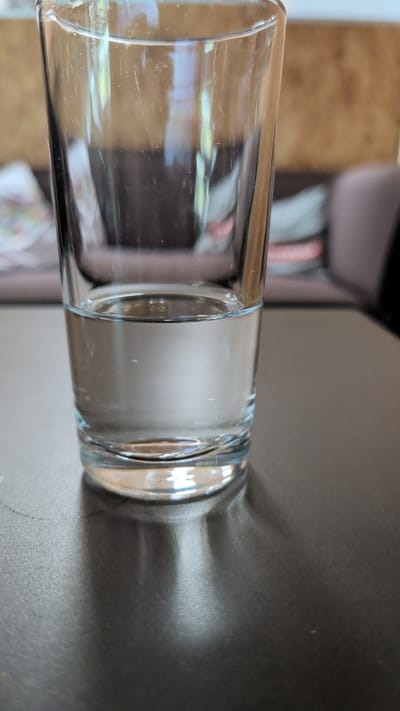 The media file is a photograph of a clear glass tumbler filled with a clear liquid, likely water. The glass is positioned horizontally and rests on a dark, textured surface, possibly a table. The tumbler is cylindrical with a thick base and a rim that appears slightly thicker than the body of the glass. The liquid inside fills approximately half of the glass, and the surface of the liquid is smooth.

To the left of the glass, a blurred background shows what appears to be furniture, with wooden tones and hints of fabric in muted colors. The lighting in the image creates a shadow of the glass on the dark surface to its right, indicating a light source from the left or above. The scene is likely indoors, given the controlled lighting and background elements. There is no visible text in the image. The overall impression is one of simplicity and focus on the object itself, with no discernible activity or specific event taking place. The image has a slightly grainy texture, suggesting it was taken with a digital camera or smartphone. The orientation of the photograph is unusual, with the glass laid on its side.