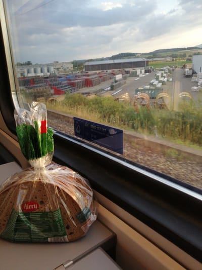 The image is a close-up shot from inside a train, looking out the window. In the foreground, a loaf of bread wrapped in clear plastic is on a tray table. The bread has a label with the brand "harry" and nutritional information. A plastic bag with green leafy vegetables and a red tag is also visible, attached to the bread packaging.

Outside the window, a train track runs parallel to the scene. Beyond the tracks, there is a large industrial area with multiple buildings, stacked pallets of goods, and what appear to be large spools or reels. The landscape in the background shows rolling hills and fields, suggesting a semi-rural or suburban setting. The sky is partly cloudy. A blue sticker with "SBB Police, Police CFF, Police FPS" and a phone number is affixed to the window. The date "07.07.26." is visible on the bread packaging. The location is identified as Eclépens, Switzerland.