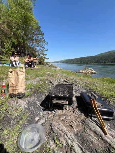 This outdoor scene captures a leisure gathering by a wide river in Sliznevo, Russia, on a bright, sunny day. A vast blue sky with faint contrails dominates the top, while forested mountains line the far bank of the river, which flows prominently on the left side of the frame. The near shore features rocky terrain interspersed with patches of green grass and scattered evergreen trees.
Two individuals are visible on the right. One person, wearing a black top and a black hat, is crouching near some trees, looking towards the river. Another person, dressed in a white top and dark bottoms, wearing sunglasses, is sitting on a light blue mat, holding what appears to be a small child or infant in their lap.
The primary activity is a barbecue or picnic, indicated by two black portable Weber grills on the rocky ground. One grill has its lid open, revealing charcoal embers, while the other's lid is closed with wooden tongs resting on top. A large brown paper bag labeled "Уголь древесный" (Charcoal wood) rests near the sitting person. Nearby, a red-capped bottle and another bottle with a yellow label, bearing the Russian text "Средство для розжига" (Lighter fluid) and a "легковоспламеняющееся" (flammable) warning, are visible. A clear plastic container lies on the ground.
The clear sky and high sun suggest it is mid-day, creating a pleasant and relaxed atmosphere for an outdoor meal by the water.