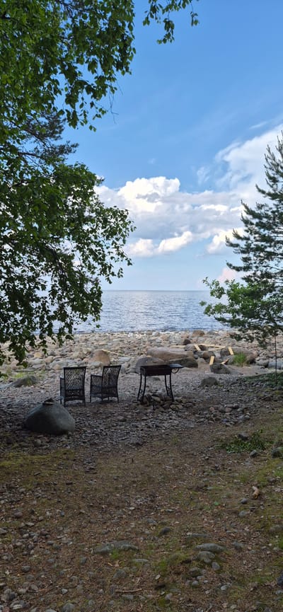 In a natural setting near the city of Motornoe, Russia, a tranquil scene unfolds by a body of water. The foreground is a stony ground with scattered pine needles and dry leaves. Two dark wicker chairs are placed on the pebbles, facing a portable barbecue grill that stands on a low pile of stones. To the left, a large, gray boulder is prominent. In the middle ground, a rocky shoreline meets the calm, rippling water of a large lake or sea. The water's surface reflects the bright, cloudy sky. Framing the scene are lush green trees, with their branches and leaves reaching into the frame from the left and right sides, adding a sense of enclosure and natural beauty. The sky is a clear blue, punctuated by large, white, cumulus clouds.