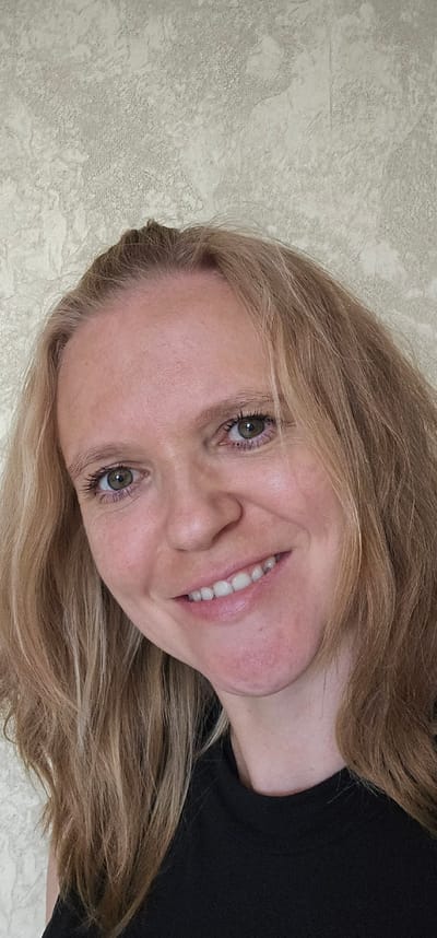 The image is a close-up, vertically oriented selfie of a blonde woman with green eyes and fair skin, smiling. She is wearing a black top. The background is a textured, light-colored wall. The image appears to have been taken indoors. There is no information available about the location in the city of Овощной, Russia.