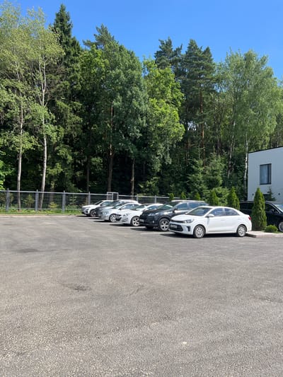 A white sedan, a dark SUV, a silver sedan, and two other silver sedans are parked in a lot. The lot is paved with asphalt and is bordered by a metal fence and a forest. A modern white building with a dark-trimmed window is visible to the right of the parking lot. The sky is clear and blue. The location is Минзаг, Russia.