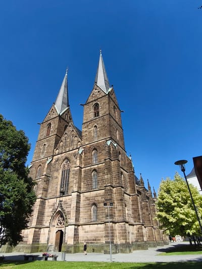 This media file shows a low-angle view of a large, Gothic-style stone church with two prominent spires against a clear, bright blue sky. A large green tree is visible to the left of the church, and a smaller tree with light green foliage is on the right. The sun is shining brightly, casting shadows and highlighting the texture of the stone.
In the foreground, on the paved area in front of the church, two people are visible. One person is lying down on a park bench, seemingly relaxing, while another person stands nearby. There is also a lamppost and some greenery. The overall scene suggests a peaceful daytime setting. The architecture of the church indicates it is likely an old and significant building, possibly a cathedral or a major parish church. The bright sunlight and clear sky suggest it is a pleasant day, likely midday or early afternoon, given the strong shadows. The presence of a park bench and people enjoying the outdoors indicates this is a public space, possibly a town square or a park adjacent to the church. There is no visible text in the image.