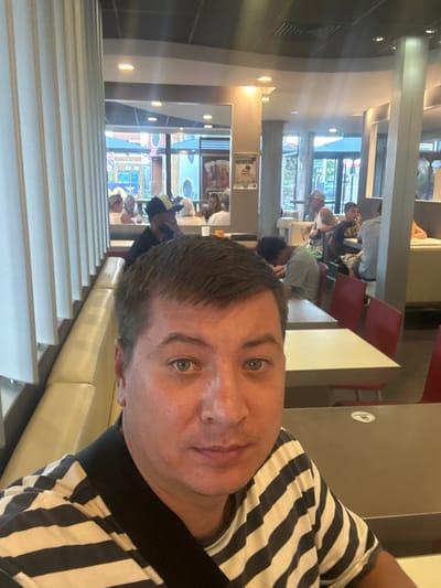 The image depicts a man in the foreground, taking a selfie in what appears to be a McDonald's restaurant in Narbonne, France. He is wearing a black and white striped shirt and has a black strap across his chest, possibly from a bag. His right eye is visible, and he is looking towards the camera with a neutral expression.
In the background, other patrons are seated at tables, some eating and others conversing. The restaurant's interior is characterized by booth seating, tables, and pillars. Sunlight streams in through windows, illuminating the space. The overall atmosphere suggests a casual dining environment. The presence of the distinctive golden arches logo on the exterior, visible through the windows, confirms the location as a McDonald's.