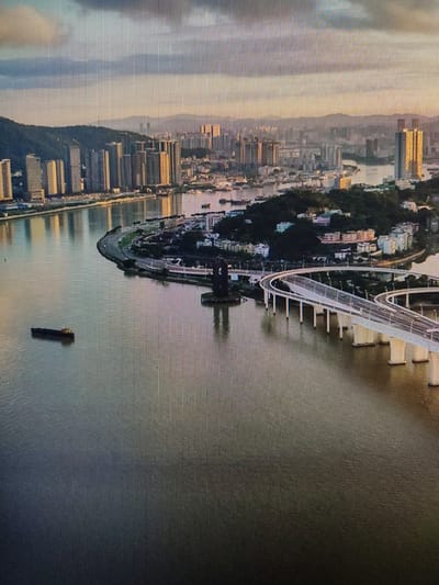 This is an aerial, medium-shot, high-angle photograph captured on a computer screen, showing a city skyline bordering a large body of water, possibly a bay or river. The scene is bathed in the soft, warm light of either sunrise or sunset, casting a golden hue on the tall buildings and the water's surface.
In the foreground, a dark-colored barge is navigating the water. To the right, a prominent, modern bridge with multiple lanes and elevated roadways curves gracefully over the water, connecting different parts of the cityscape. The bridge's white structure stands in stark contrast to the darker water and surrounding land.
The mid-ground reveals a densely populated urban area with numerous high-rise buildings, many of which are contemporary in design and reflect the ambient light. Some of these buildings are clustered on hillsides, suggesting a varied topography. The water in this area appears calmer, with reflections of the buildings visible.
The background features a hazy, mountainous landscape further in the distance, with more indistinct city structures scattered between the hills. The sky is partially obscured by clouds, with streaks of light breaking through, indicating dynamic weather conditions or the time of day.
The image lacks any discernible human figures. The overall impression is one of an expansive coastal or riverside metropolis during a transitional period of light, highlighting urban development and infrastructure. The photograph's pixelated quality suggests it has been taken from a display screen. The prompt's mention of "Bielany Wrocławskie, Poland" is not supported by any visual cues within the image itself.