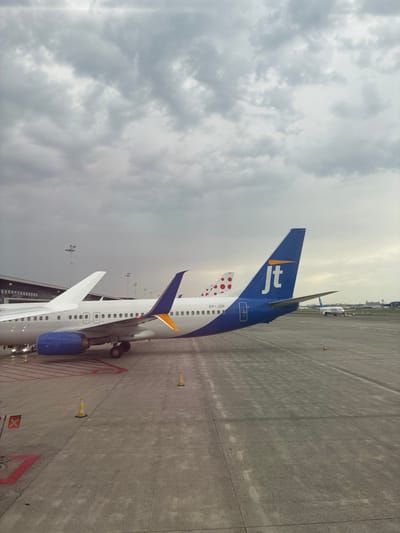 An overcast sky looms over an airport tarmac at Brussels Airport in Steenokkerzeel, Belgium. In the foreground, two airplanes are parked. The nearest aircraft has a white fuselage, a blue engine cowling, and a tail fin adorned with a blue "jt" logo and an orange stripe. The registration "OY-JZP" is visible on its fuselage. Behind this plane, another aircraft's tail fin, decorated with red polka dots, is partially visible. Further in the background, a third airplane with "Air Europa" written on its side is taxiing on the tarmac. The ground is concrete, marked with red and white lines and dotted with traffic cones. Airport buildings and lighting poles are visible in the distance.