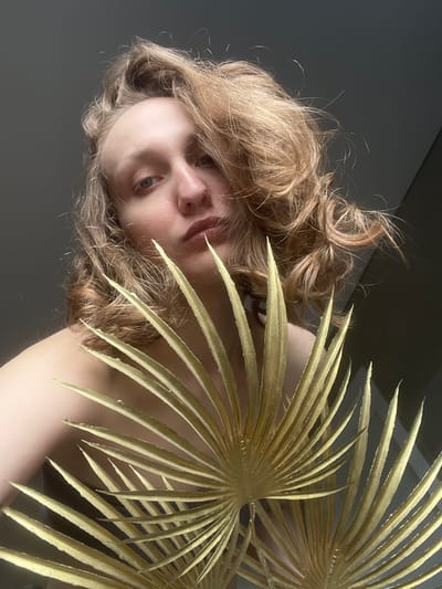 A low-angle shot depicts a person with blonde, wavy hair partially obscured by a golden palm frond. The person's face is visible, with a clear view of their nose, lips, and one eye. The background is a mix of gray and neutral tones, with an indication of a ceiling line. The lighting appears diffused and even. The image does not definitively provide any information about the city of Podshivalovo, Russia.
