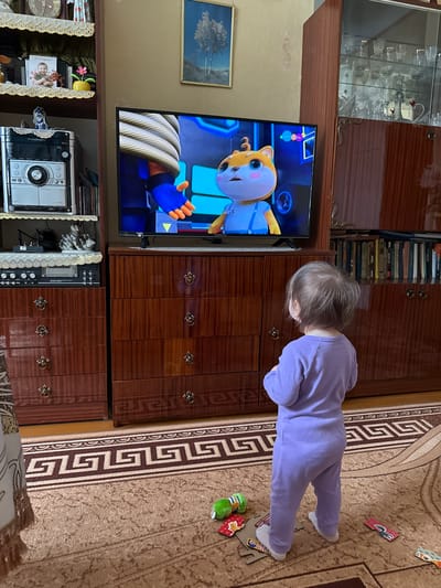 A child in a lavender jumpsuit is watching a cartoon on a television set. The TV is set inside a brown wooden cabinet. To the left of the TV, is a clear glass cabinet filled with glassware. There is a picture hanging on the wall behind the TV. Below the TV is a stereo system. The child is standing on a brown carpet with a geometric pattern and puzzle pieces scattered on the floor. The location is likely Vyshegor, Russia, based on the provided information.