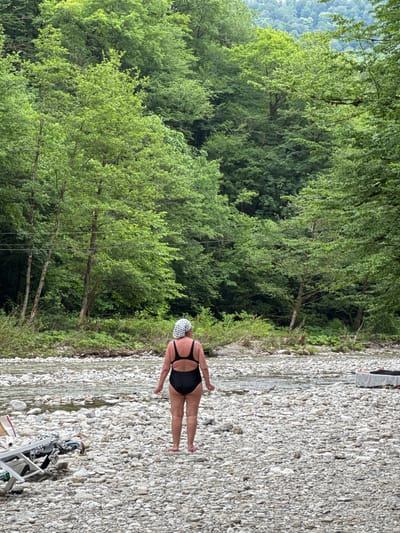 A person stands on a rocky riverbed, facing away from the camera, towards a shallow, clear river. The person is wearing a black one-piece swimsuit and a white patterned headscarf. The riverbed is covered in smooth, grey and white stones of various sizes. The river is shallow, with visible stones beneath the surface. On the left side of the image, there are some pieces of furniture and bags on the shore. The background is dominated by a dense, lush green forest that rises up a hill. Power lines are visible strung between two trees on the left side of the frame. The sky is overcast, creating a soft, diffused light. The scene appears to be a natural, undeveloped area, likely a riverbank in a forested region. The location is identified as Khleborob, Russia.