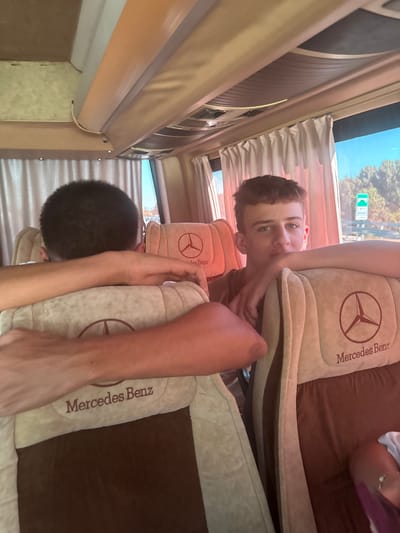 The image depicts the interior of a bus, as evidenced by the rows of seats and the overhead compartments. Two young males are visible. One, in the foreground and facing away from the camera, has dark, short hair and his arms are resting on the back of a seat. The second male, in the mid-ground and facing the camera, has light brown hair and is looking directly at the viewer with his right arm resting on the seat back. The seats are upholstered in a light beige fabric with dark brown accents and feature the Mercedes-Benz logo. The environment outside the bus windows appears to be a sunny day with trees and a road sign visible, suggesting a location in Albania, possibly near Fushë-Krujë, though this cannot be definitively confirmed from the image alone. The lighting suggests it is daytime.