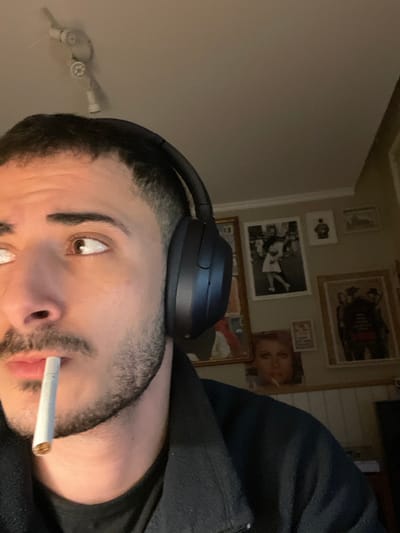 A close-up, rotated image captures a man from a low angle, looking slightly upwards and to the side. He has short, dark hair with a fade, a beard, and brown eyes. He is wearing dark over-ear headphones and has an unlit cigarette held between his lips. A dark, possibly black, hooded garment is visible on his shoulder.
The background features a light-colored wall decorated with several framed pictures and posters. These include a large, dark framed piece, a smaller framed image below it, a medium-sized black and white photograph of multiple figures (possibly an event or sports scene), and a larger framed piece with a reflective surface. Notably, an unframed or loosely framed image of a woman with a cigarette in her mouth is visible on the wall, mirroring the man's action. A part of a light fixture or small ceiling fan is also seen on the upper left wall. The indoor lighting appears somewhat dim. No text is visible in the image. The scene takes place indoors, potentially in Ganei Tikva, Israel.