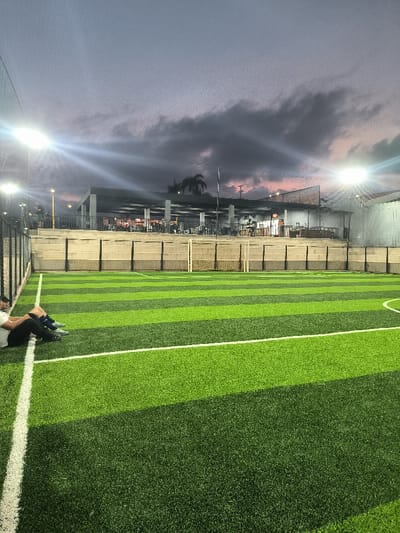 The image captures a vibrant artificial turf soccer field at dusk or early evening, illuminated by bright floodlights. The field features distinct alternating stripes of dark green and light green artificial grass, with crisp white lines marking the boundaries and a partial arc on the right side, likely part of a penalty area. A small white soccer goal stands in the background, slightly to the right of the center.
Along the left edge of the field, a person is seated on the white sideline, leaning against what appears to be a protective fence or wall. They are wearing a white t-shirt and dark shorts or pants, with athletic shoes. Their posture suggests rest or observation.
The field is enclosed by a tall dark fence, visible on the left and extending upwards, with some mesh netting above it. Beyond the fence, in the background, there is an elevated structure with a roof supported by pillars, resembling a viewing stand or a commercial building associated with the sports complex. Several individuals can be faintly seen on this upper level. In the distance, a prominent palm tree and other vegetation are silhouetted against the sky, along with utility poles and wires.
The sky above is a notable feature, displaying the soft hues of twilight. Dark grey clouds stretch across the frame, interspersed with lighter purplish-pink tones, indicating the sun has either just set or is about to rise. The artificial floodlights on tall poles, particularly one on the top-left and another on the top-right, cast strong beams of light across the field and upward, creating significant glare. The overall lighting suggests that play or practice is either ongoing or about to commence/conclude as natural light fades. There is no discernible text visible in the image.
