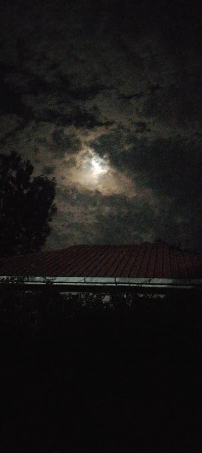 This image depicts a nighttime scene in South Kasipul ward, Kenya. The sky, occupying the upper half of the frame, is overcast with dark, textured clouds. A bright, diffused light source, consistent with the moon, is visible through a break in the clouds towards the upper-center, illuminating portions of the cloud cover. To the left, a dark, indistinct silhouette of a tree is partially visible against the cloudy sky. Below the horizon line formed by the clouds, the angled red-brown tiled roof of a structure is visible, extending across the mid-ground. Below the roof, a light-colored gutter runs horizontally. The lower portion of the image is predominantly dark, indicating minimal or no artificial light sources, with faint suggestions of foliage immediately below the gutter. No people, subjects, actions, or interactions are discernible.