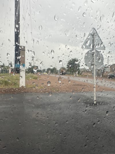 The image shows a view through a rain-streaked window, likely from inside a vehicle. The foreground is dominated by the texture of water droplets on the glass, obscuring a clear view of the outside. Beyond the window, a road is visible, appearing wet and reflecting the overcast sky. There are some buildings and vegetation in the background, suggesting an outdoor, possibly rural or suburban, environment. A traffic sign, a triangular yield sign on a post, is also discernible through the rain. The overall atmosphere is grey and damp, indicative of rainy weather. The location is identified as Sarymoldayev Rural District, Kazakhstan, which aligns with the observed scenery of a road, structures, and vegetation under a cloudy sky.