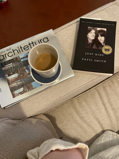 The image shows a top-down view of a beige upholstered couch with two books and a coffee mug on it. One book is open, displaying an architectural photograph of ancient ruins. The second book, titled "JUST KIDS" by PATTI SMITH, is closed and has a "NATIONAL BOOK AWARD WINNER" seal on its cover. A white ceramic coffee mug with a light brown liquid, presumably coffee, sits on a dark blue coaster on top of the open book. The overall scene suggests a moment of relaxation and reading. There is no indication of Piacenza, Italy, or any specific city.