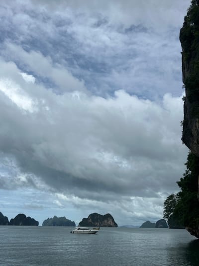 The image depicts a scenic view of Phang Nga Bay, Tambon Krasom, Thailand, under a cloudy sky. The foreground is dominated by a vast expanse of calm, grey water, with a gentle ripple across its surface. In the middle ground, a white speed boat with a yellow stripe is traveling from left to right across the frame, its wake subtly visible. In the background, a series of lush, green limestone karsts rise dramatically from the sea, their peaks softened by distance and haze. The sky is filled with a dramatic display of heavy, grey clouds, interspersed with patches of lighter grey and hints of blue sky peeking through. To the right of the frame, a large, dark cliff face, covered in dense green vegetation, juts out into the sea, partially obscuring the view of the karsts and the sky. The overall atmosphere is moody and serene, suggesting an overcast day in a tropical marine environment.