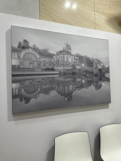The image displays a framed black and white photograph hanging on a textured white wall. The photograph is oriented vertically and appears to be a reflection of a city street scene. The top portion of the photograph shows an inverted view of buildings with windows, rooftops, and trees. There are streetlights visible, suggesting it might be dusk or nighttime. The lower portion of the photograph is the reflection of this scene, creating a symmetrical effect. The architecture suggests a European city. To the left of the photograph, a section of a floor with a travertine tile pattern is visible, and to the right of the photograph, two curved white objects, possibly decorative elements or light fixtures, are mounted on the wall. The overall mood of the photograph is somewhat surreal due to the mirrored reflection. There is no visible text in the image.