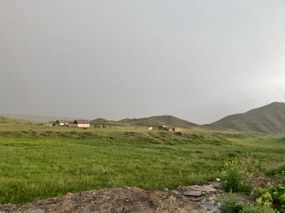 The image depicts a rural landscape in Kara-Oi, Kyrgyzstan, under an overcast sky. In the foreground, a muddy patch of ground with some scattered debris is visible, bordered by a low wire fence. Beyond the fence, a vast, green, grassy field stretches towards a cluster of small houses. These houses are low-lying structures with tiled or metal roofs in shades of red and yellow. Some of the houses appear to be made of brick or mud. In the middle ground, rolling hills rise, sparsely dotted with more buildings and occasional trees. The sky is a uniform, light gray, suggesting a cloudy or potentially rainy day. The overall impression is one of a simple, undeveloped rural settlement set against a backdrop of natural, hilly terrain.