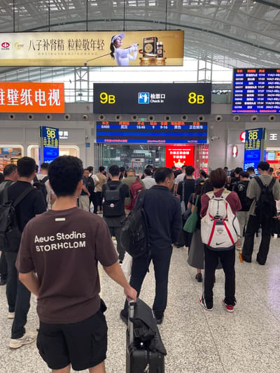 This image captures a busy scene at a train station in Longhua District, China. A crowd of people, dressed in casual attire, are standing or walking, many with luggage. The environment is indoors with a high ceiling and fluorescent lighting. Large digital displays are prominently featured, showing train information such as departure times, destinations, and gate numbers (e.g., "9B", "8B"). One display indicates "Check in" with a pictogram. Advertisements are visible on the walls, including one with Chinese characters and an image of a woman playing golf. The floor is tiled with a speckled pattern. A person in the foreground, facing away from the camera, wears a brown t-shirt with white text reading "Aeuc Stodins STORHCLOM" and black shorts, holding a rolling suitcase. Another person nearby is wearing a white and red backpack. The overall atmosphere suggests transit activity and waiting for trains.