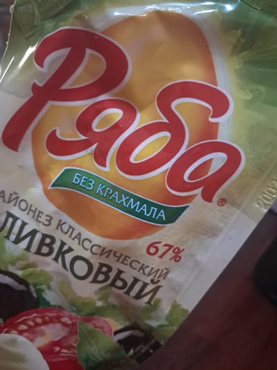The image shows a close-up of a food product package, likely mayonnaise, with Russian text. The brand name appears to be "Ряба" (Ryaba). There is text that reads "БЕЗ КРАХМАЛА" (Bez krakhmala), meaning "without starch." Below this, it says "МАЙОНЕЗ КЛАССИЧЕСКИЙ" (Mayonez klassicheskiy), meaning "classic mayonnaise," and then "ЛИВКОВЫЙ" (Livkovyy) which seems to be part of the product name or description. A percentage "67%" is also visible. The packaging features images of fresh vegetables such as tomatoes and olives. The background is a light green and yellow gradient. The image quality is poor, with a blurry foreground and a slightly out-of-focus background. There is no discernible urban or rural environment in Chobruchi, Moldova, as the focus is solely on the product packaging. No people are visible in the image.